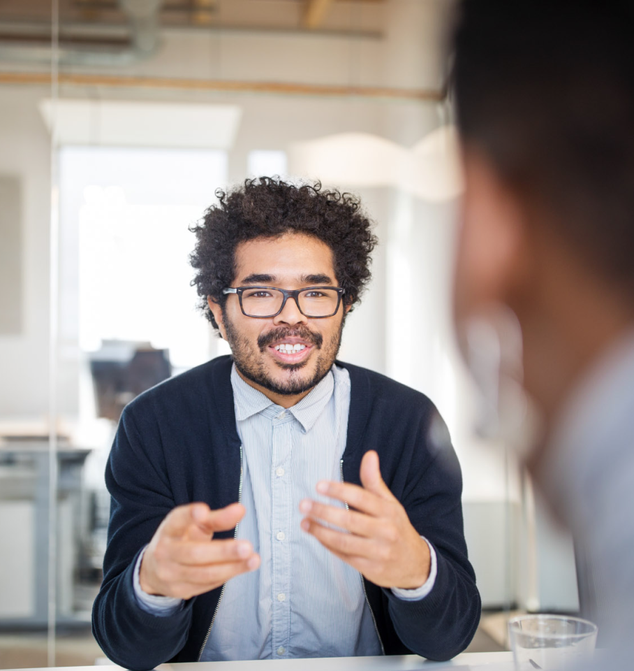 Image is of a young male healthcare professional in an office having a conversation with a colleague.